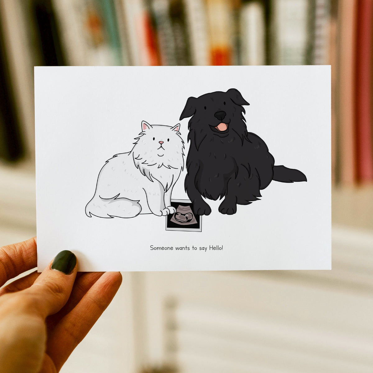 A person holding a horizontal white announcement card that features a hand-drawn illustration of a fluffy white Persian cat and a long-haired black dog sitting next to a baby ultrasound photo, with the text "Someone wants to say Hello!" printed at the bottom.