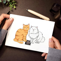 A person holding a horizontal white card over a dark desk, featuring a hand-drawn illustration of a ginger tabby cat and a white Persian cat sitting together next to a baby ultrasound photo.