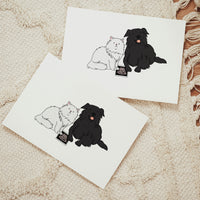 Two horizontal white announcement cards arranged on a woven cream rug, displaying identical hand-drawn illustrations of a fluffy white cat and a long-haired black dog sitting next to a baby ultrasound photo.