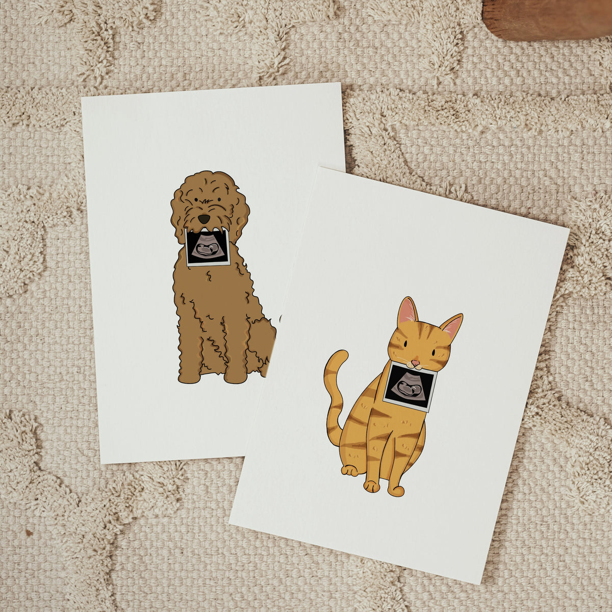 Two vertical white cards overlapping on a textured cream carpet; one displays a hand-drawn brown doodle dog holding a baby ultrasound photo, and the other shows an orange tabby cat holding a baby ultrasound photo.