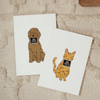 Two vertical white cards overlapping on a textured cream carpet; one displays a hand-drawn brown doodle dog holding a baby ultrasound photo, and the other shows an orange tabby cat holding a baby ultrasound photo.
