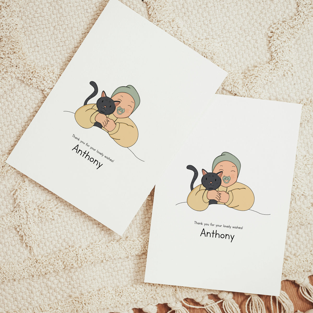 Two vertical personalized birth announcement cards displayed on a beige woven rug. The realistic vector illustration features a smiling baby wearing a sage green beanie and a yellow romper, happily hugging a sleek black cat. Below the art, the printed text reads "Thank you for your lovely wishes! Anthony"
