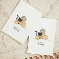 Two vertical personalized birth announcement cards displayed on a beige woven rug. The realistic vector illustration features a smiling baby wearing a sage green beanie and a yellow romper, happily hugging a sleek black cat. Below the art, the printed text reads "Thank you for your lovely wishes! Anthony"