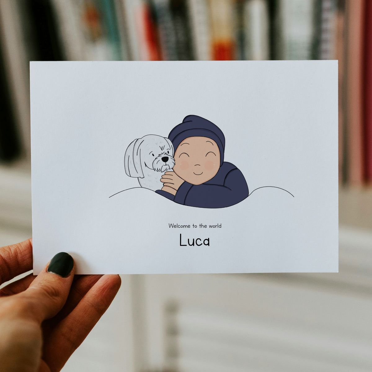 A close-up view of a hand holding a horizontal custom baby card. The minimalist vector drawing features a smiling baby in a dark navy blue romper and matching beanie, hugging a fluffy white Maltese puppy. The printed text below the illustration reads "Welcome to the world Luca".