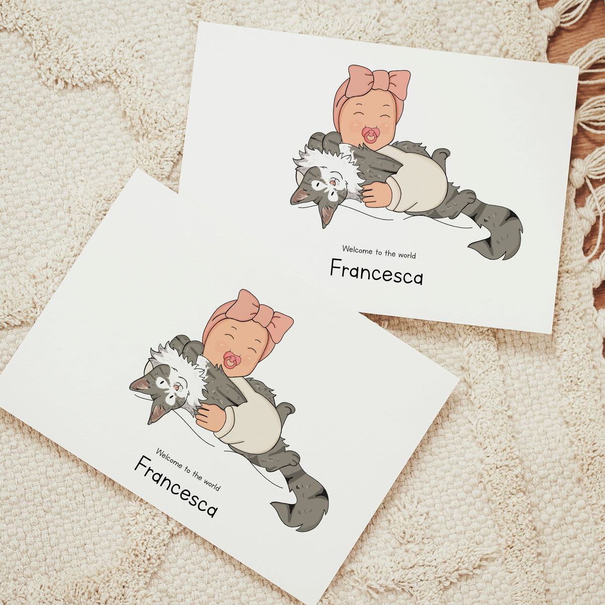 Two custom horizontal birth announcement cards on a textured cream rug. The hand-drawn vector illustration shows a laughing baby wearing a pink bow headband and a cream romper, holding a large, fluffy gray and white tabby cat on its back. The text below reads "Welcome to the world Francesca"