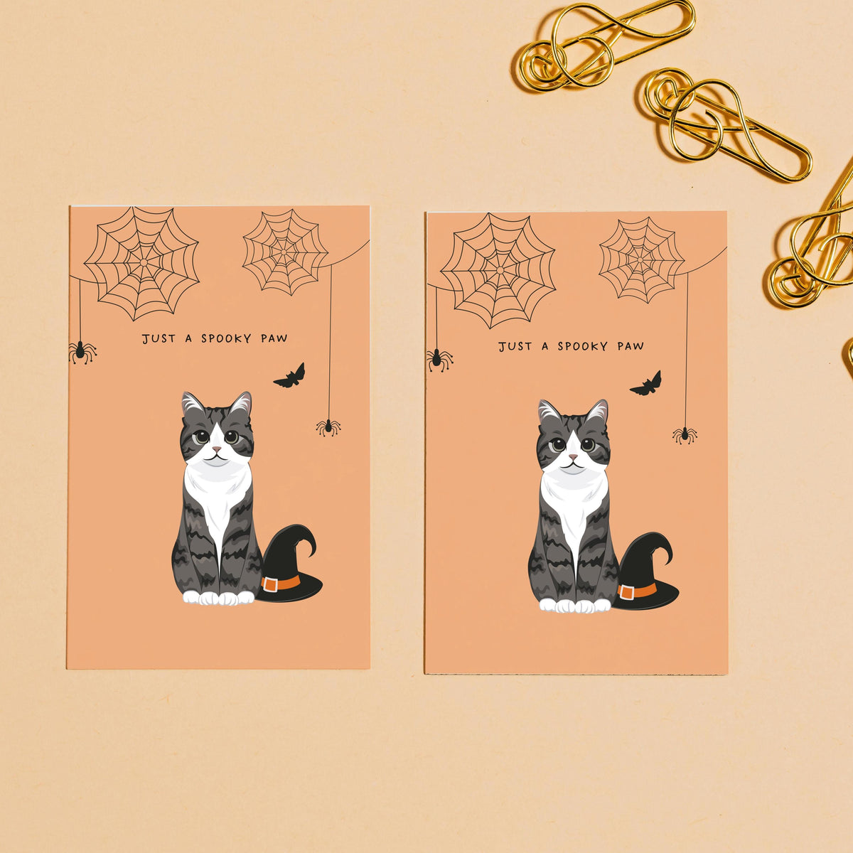 Flat lay of two side-by-side custom greeting cards on a beige background with gold paperclips. Each card features a cartoon caricature portrait of a grey and white tabby cat next to a black witch hat under hanging spiderwebs, designed as a Personalized Pet Portrait Halloween Card with the text "JUST A SPOOKY PAW".