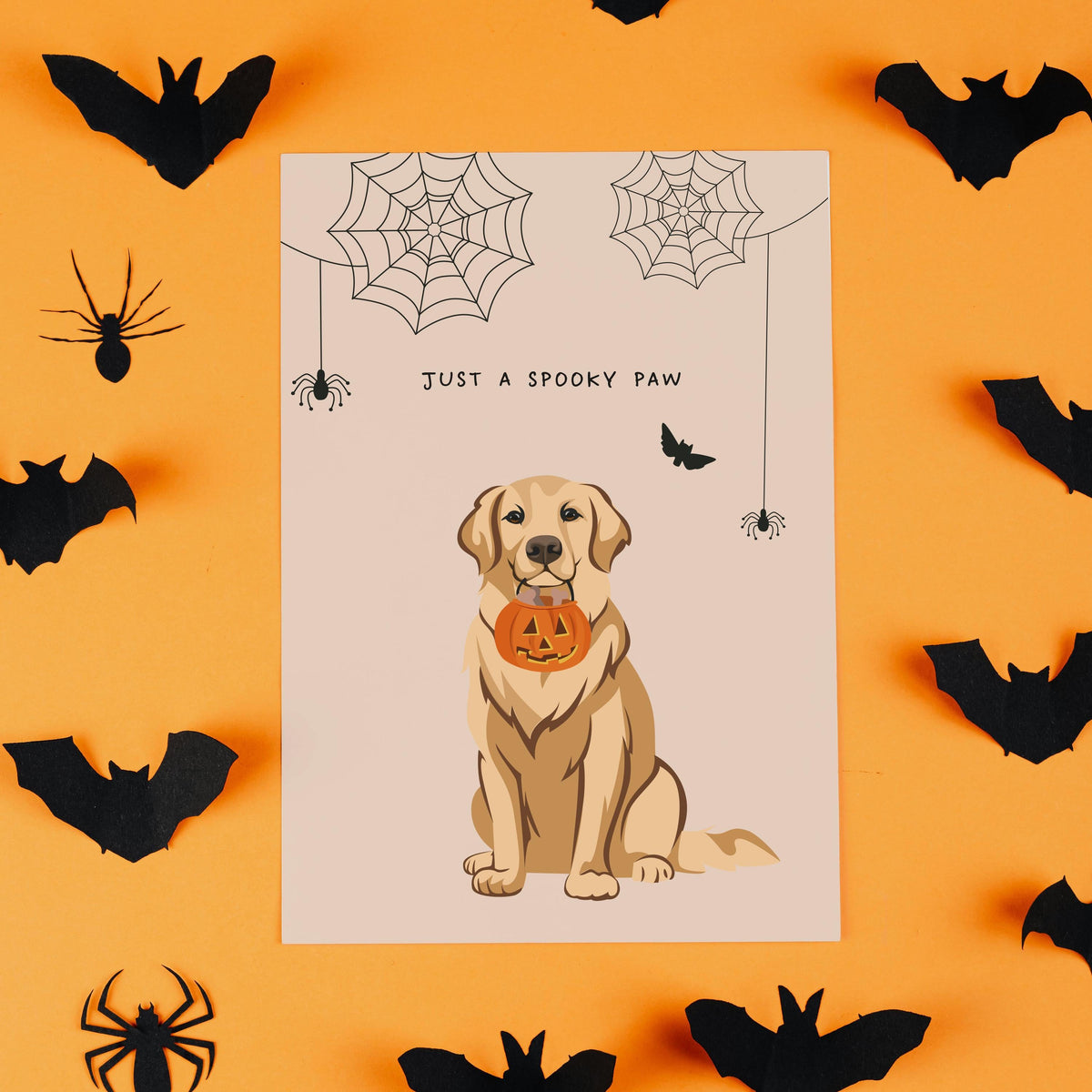 Flat lay of a custom holiday card surrounded by black paper cutout bats and spiders on a vibrant orange background. The card displays a realistic illustration of a golden retriever dog holding a jack-o'-lantern pumpkin candy bucket, customized as a Personalized Pet Portrait Halloween Card with the text "JUST A SPOOKY PAW"