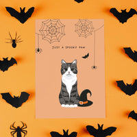 Top-down view of a single custom greeting card centered among black paper bats and spiders on a bold orange surface. The design features a caricature cartoon portrait of a grey tabby cat next to a black witch's hat, acting as a Personalized Pet Portrait Halloween Card with the caption "JUST A SPOOKY PAW".