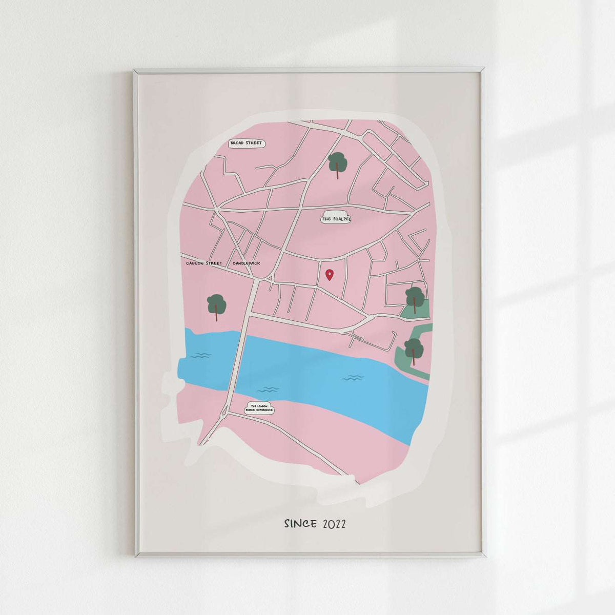 A minimalist personalized map art print of London in pink and blue, featuring a red location pin near The Scalpel and Cannon Street. The custom map includes hand-drawn street details, a river, and 'Since 2022' text at the bottom. Framed against a white wall with soft shadows.