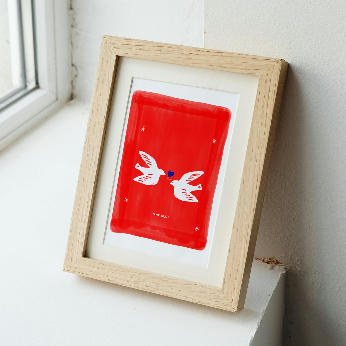 A wooden framed art print of two personalized white doves meeting over a blue heart. The artwork features a vibrant red textured background with white stars and the custom script "amour" at the bottom, showcasing an artisanal, human-led design perfect for a wedding or anniversary gift.