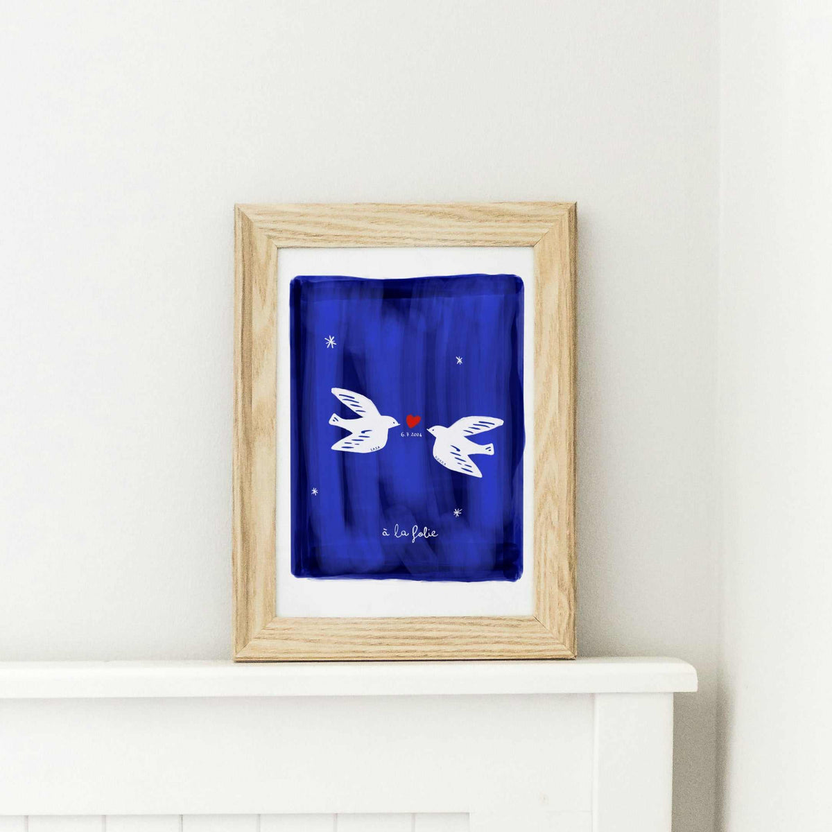 A wooden framed art print of two personalized white doves meeting over a red heart. The artwork features a deep navy blue textured background with white stars and the custom script "à la folie" at the bottom, showcasing an artisanal, human-led design perfect for a wedding or anniversary gift.