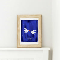 A wooden framed art print of two personalized white doves meeting over a red heart. The artwork features a deep navy blue textured background with white stars and the custom script "à la folie" at the bottom, showcasing an artisanal, human-led design perfect for a wedding or anniversary gift.