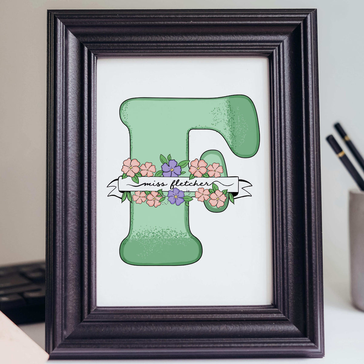 A hand-drawn personalized teacher appreciation gift featuring a large mint green letter "F" decorated with pink and purple blossoms. A white ribbon banner wraps around the letter with the name "Miss Fletcher" in elegant black script. The artwork is displayed in a classic black frame on a desk, representing a unique gift for teachers from students that feels personal even without knowing their hobbies.