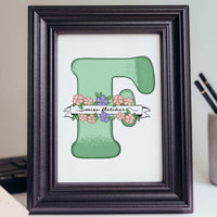 A hand-drawn personalized teacher appreciation gift featuring a large mint green letter "F" decorated with pink and purple blossoms. A white ribbon banner wraps around the letter with the name "Miss Fletcher" in elegant black script. The artwork is displayed in a classic black frame on a desk, representing a unique gift for teachers from students that feels personal even without knowing their hobbies.