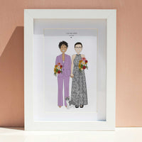 Framed custom wedding illustration portrait of a same-sex couple, one in a lavender suit and one in a grey floral dress, holding hands with their cat, displayed in a white frame against a peach wall.